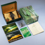 A Rolex Submariner watch is displayed with its original green presentation box, a second green box, multiple brand brochures, a warranty card, and a small accessory packet on a light blue-gray background.