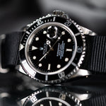 A Rolex Submariner watch with a black dial, black and silver bezel, and a black fabric strap, shown in a close-up shot with a reflective surface below.