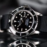 A Rolex Submariner watch with a black dial, black and silver bezel, and a black fabric strap, shown in a close-up shot with a reflective surface below.