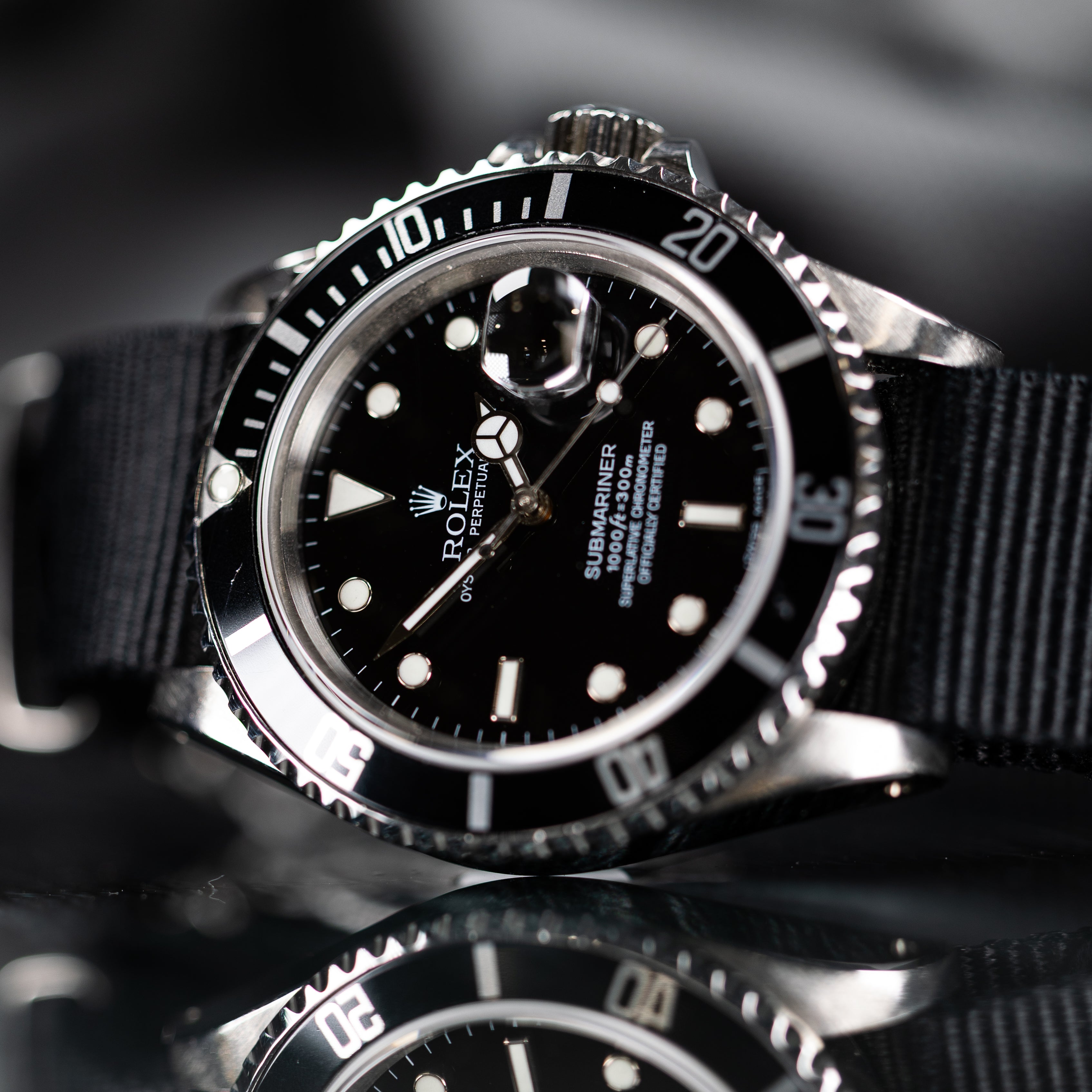 A Rolex Submariner watch with a black dial, black and silver bezel, and a black fabric strap, shown in a close-up shot with a reflective surface below.
