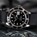 A Rolex Submariner Date watch with a black dial, black bezel, and a black nato strap, shown in a close-up shot with a reflective surface below.