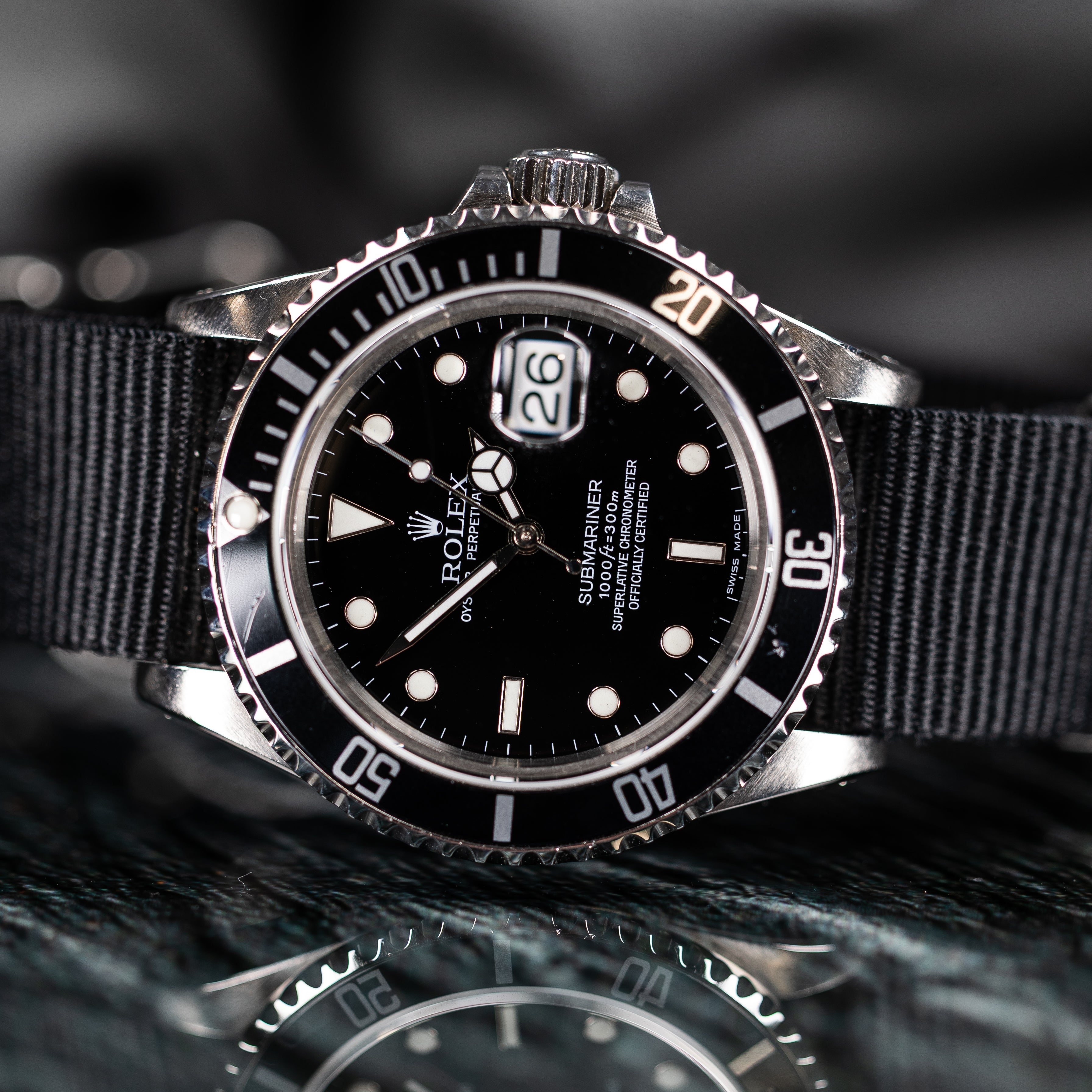 A Rolex Submariner Date watch with a black dial, black bezel, and a black nato strap, shown in a close-up shot with a reflective surface below.