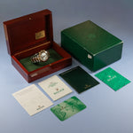 A Rolex watch displayed inside an open dark wood presentation box, alongside a green Rolex-branded outer box, manuals, cards, and a green wristlet, all arranged on a light blue studio background.
