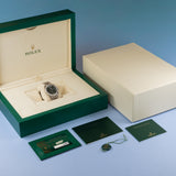 Rolex watch presentation set including an open green box with a silver-toned watch inside, a closed cream-colored outer box, and several green documentation cards on a blue gradient background.