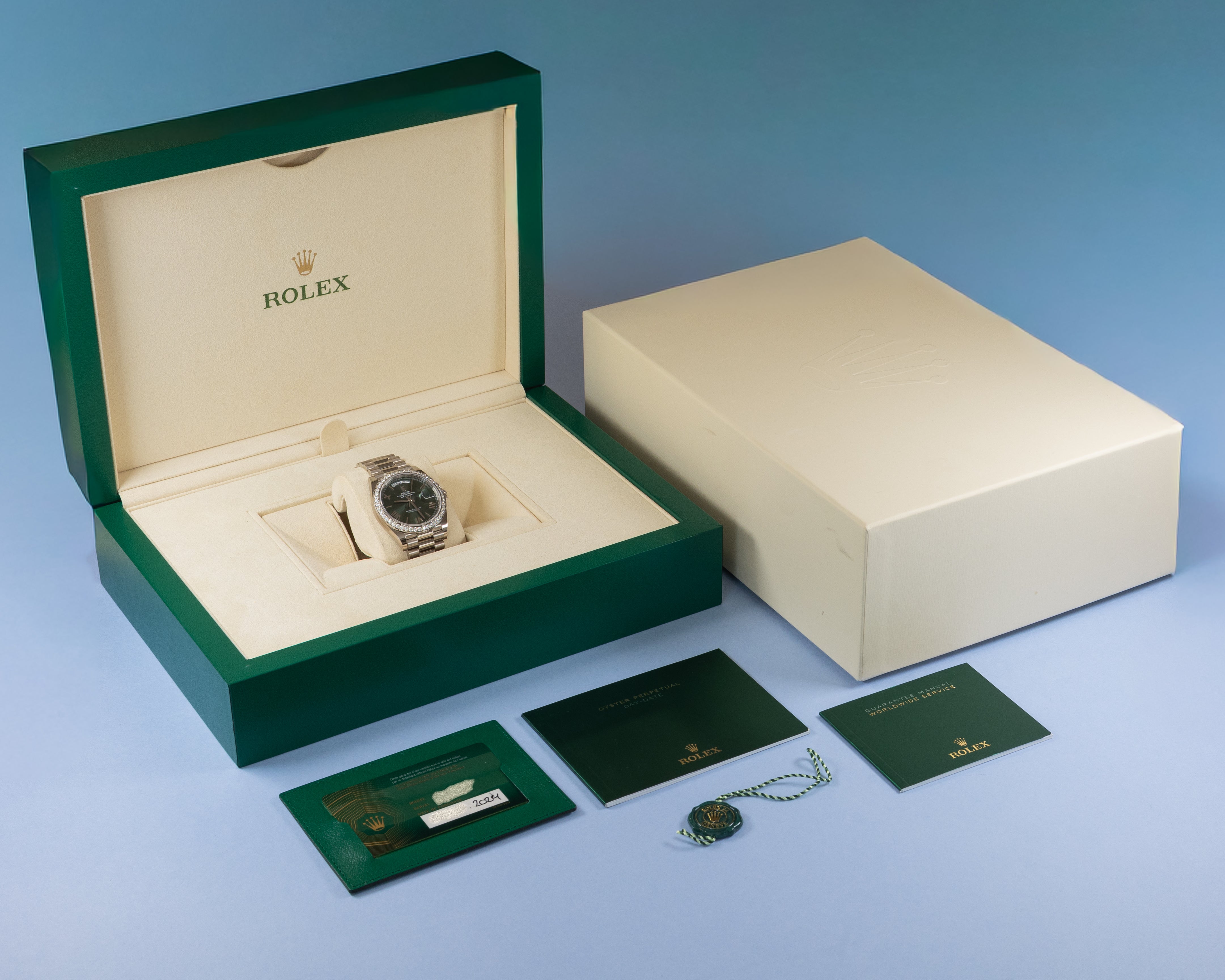 Rolex watch presentation set including an open green box with a silver-toned watch inside, a closed cream-colored outer box, and several green documentation cards on a blue gradient background.