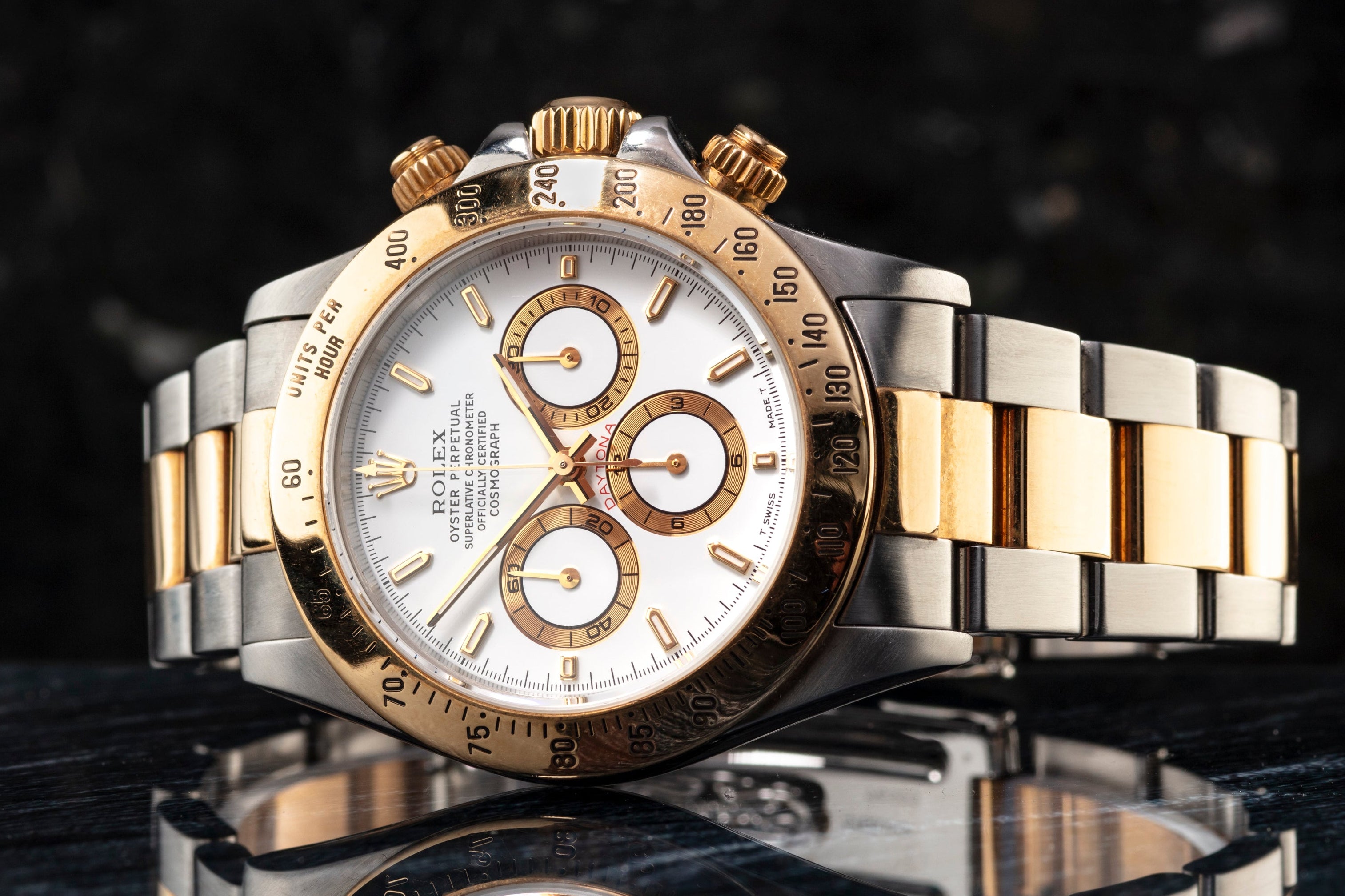 A luxury two-tone wristwatch with a silver and gold metal bracelet, a gold bezel, and a white dial, displayed on a reflective dark surface against a dark textured background.